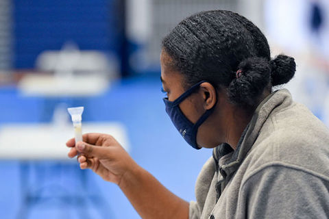 COVID-19 Disparities. Black woman with dark hair in two buns wearing a black mask and gray top holding a vial of saliva.