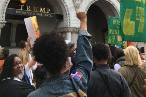 An assembled crowd of mostly women in front of Trump Hotel in Washington, D.C. One Black woman with a natural up-do, has her fist raised in the air.