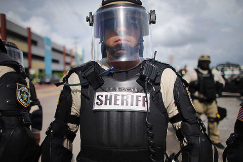 A sheriff in riot gear wears a clear face shield and has the word SHERIFF written in large letters across his bullet-proof vest.