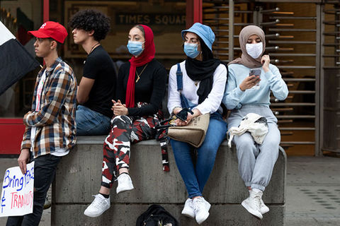 Hijabs. Three women in masks sit on a cinderblock next to two young men without masks.