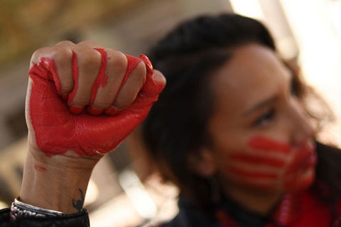 Indigenous Woman. Raised fist painted red with dark-haired woman with mouth covered in red hand in the background.