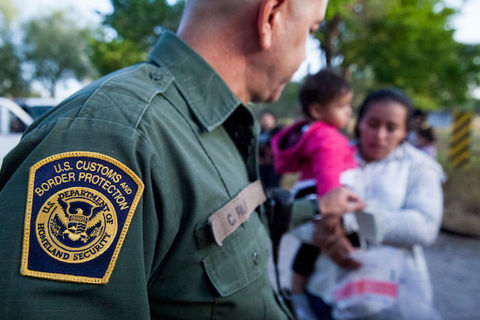Foreground shows the arm of a man in a green shirt with a patch that reads, US Customs and Border Protection. The background shows a woman holding a small child wearing a pink jacket.