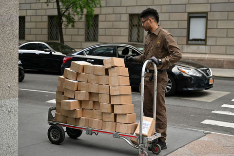 A black man wearing a brown UPS uniform pushes a dally filled with brown, boxed packaged.