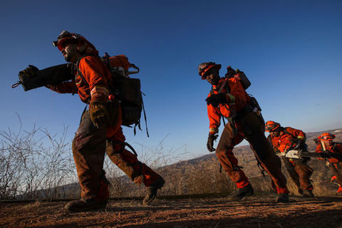 People standing outside in a line are wearing fireman uniforms and carrying firefighting equipment. 