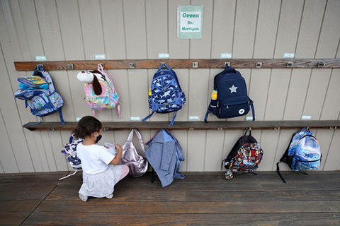 LA Schools. Small girl child kneels on floor in front of a row of back packs hanging from a wall.