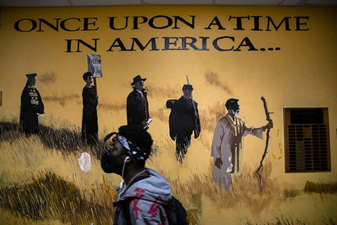 Stamford CT High School. Black male student wearing bandana, black face mask, hoodie and backpack walks by wall mural reading "once upon a time in America."