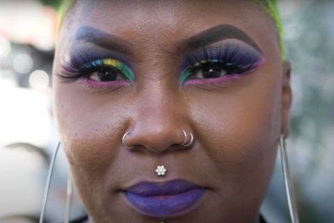 Black woman with very close cropped blonde hair, and colorful eye make up (green, yellow, purple) as well as several nose rings and purple lipstick