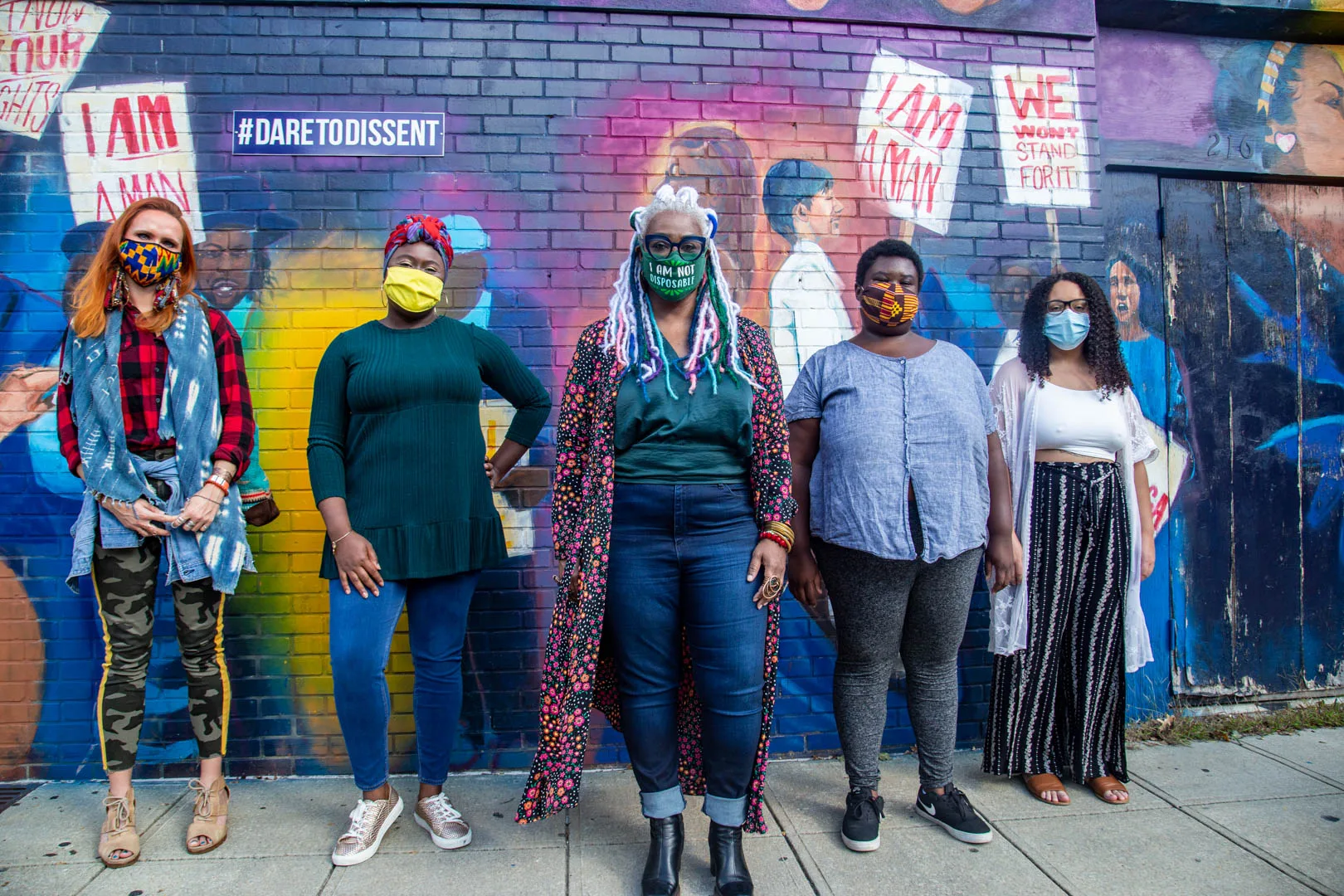 Five people in colorful clothing and face masks standing in front of a wall painted with images of people holding protest signs.